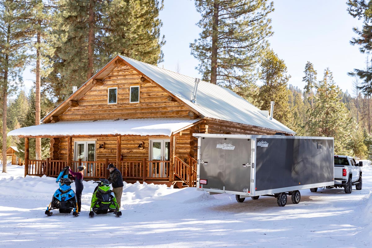 Enclosed Deckover 101" Wide Snow Grey Trailer With Snowmobiles In Front Of Cabins